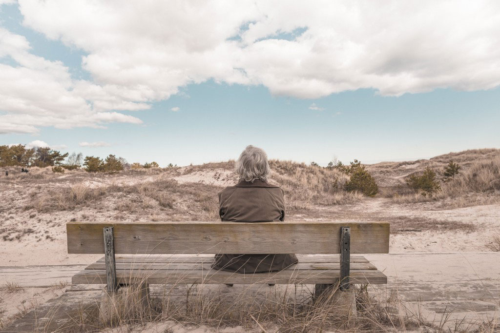 older woman sits on a beachside bench looking over the sand dunes